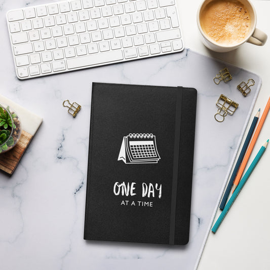 Schwarzes Notizbuch mit weißem Kalender-Motiv und dem Text: „One day at a time“