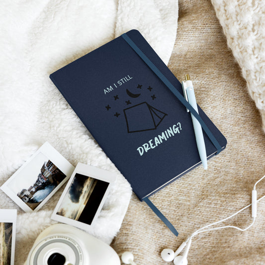 Dunkelblaues Notizbuch auf heller Decke, mit weißem und schwarzem Design. Aufdruck: „Am I still dreaming?“ unter einer Zelt- und Sternengrafik.