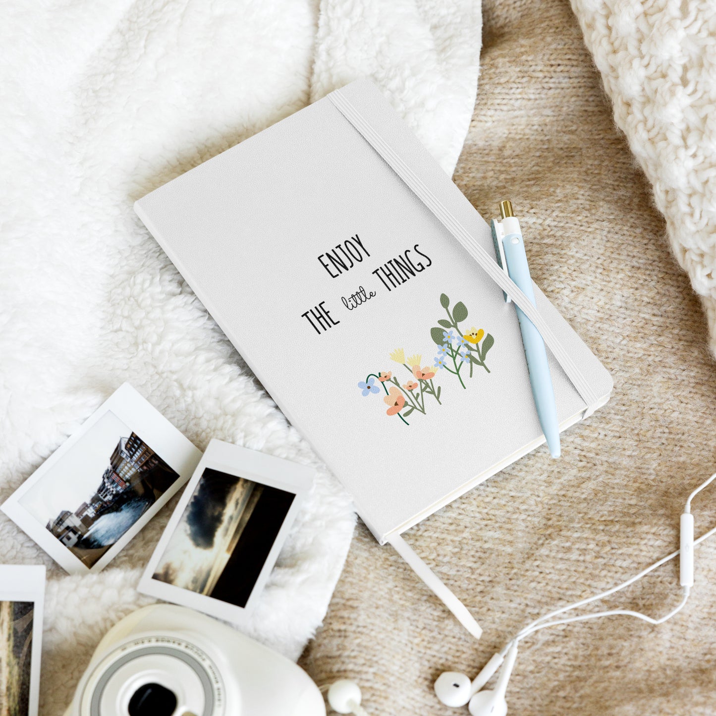 White notebook with "Enjoy the little things" and colorful flowers, on a cozy blanket with Polaroid photos and a blue pen.