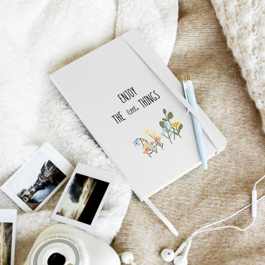 White notebook with "Enjoy the little things" and colorful flowers, on a cozy blanket with Polaroid photos and a blue pen.