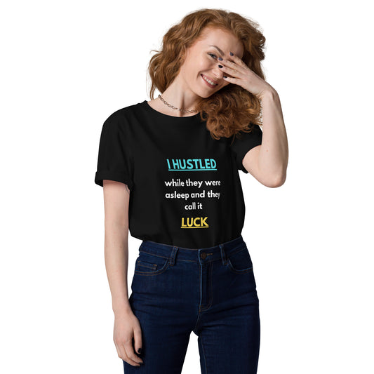 Schwarzes T-Shirt mit dem Spruch: „I hustled while they were asleep and they call it luck“, wobei „hustled“ blau und „luck“ gelb hervorgehoben ist.